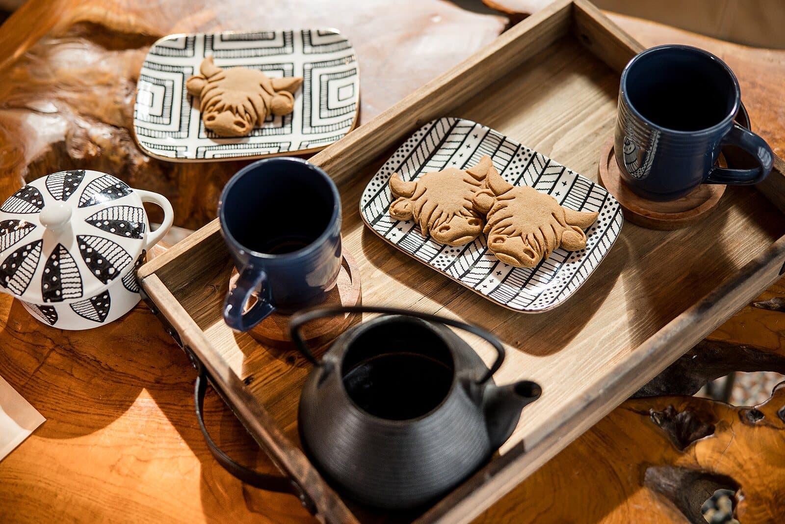 Highland cow-shaped gingerbread cookies on patterned plates with blue mugs and teapots on a wooden tray