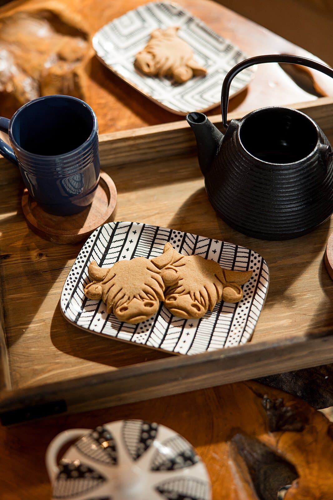 Two Highland cow-shaped gingerbread cookies on a patterned plate beside a black teapot and blue mug