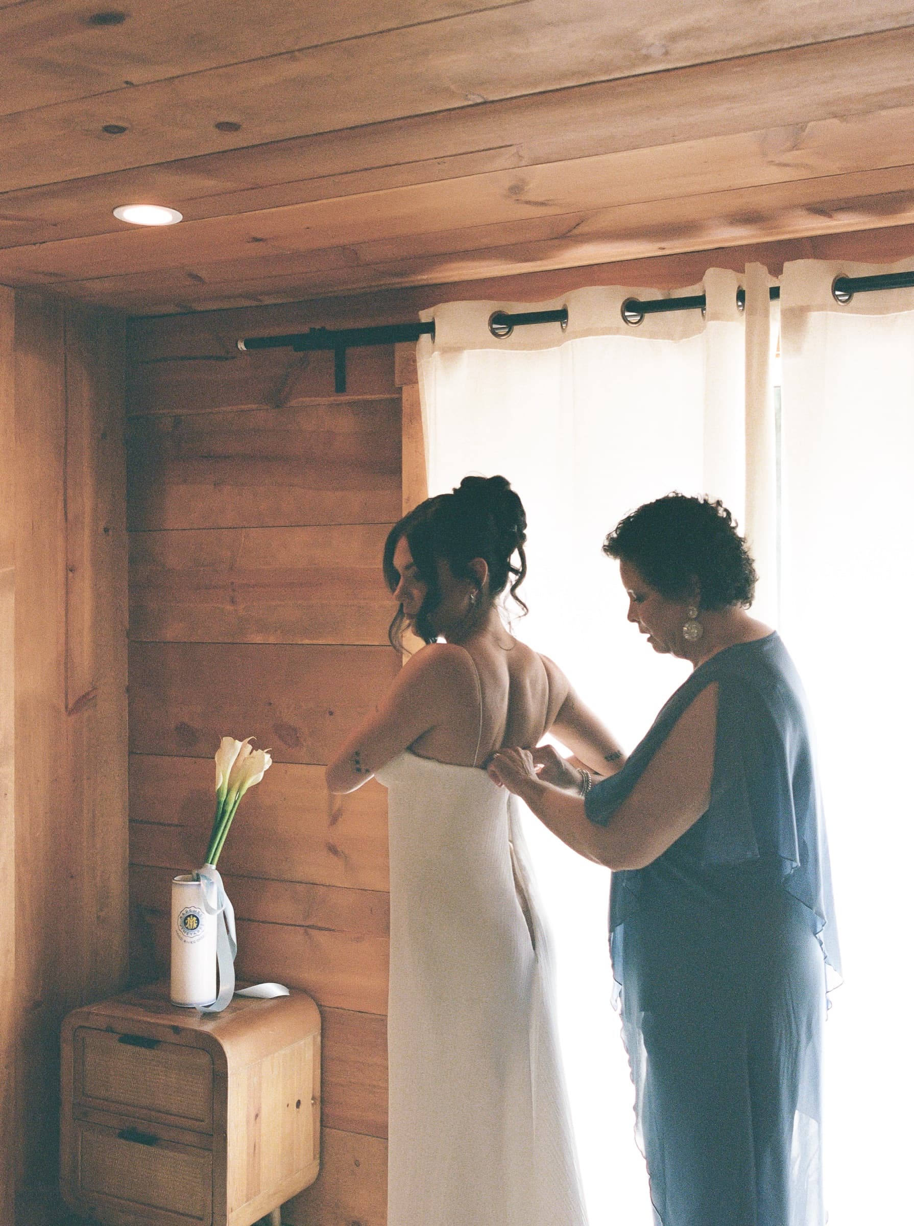An older woman fastens a bride's dress beside a window in a cedar-walled room