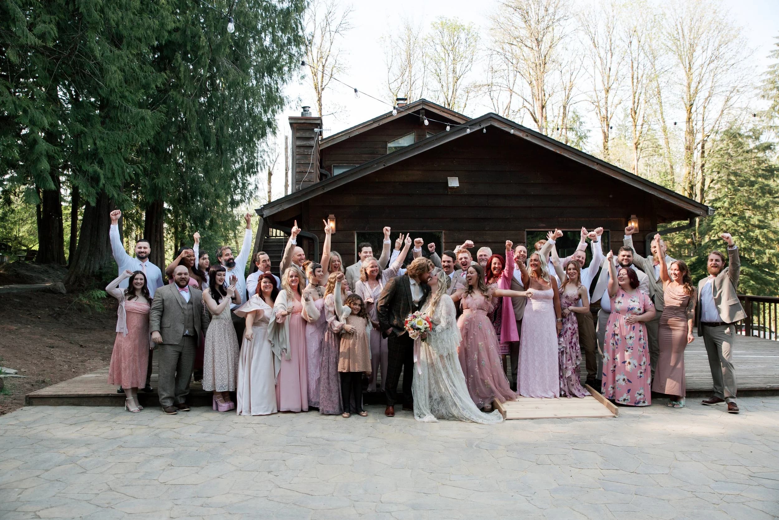 Sydney and Casey with their whole wedding party, arms raised, in front of a dark-timbered building